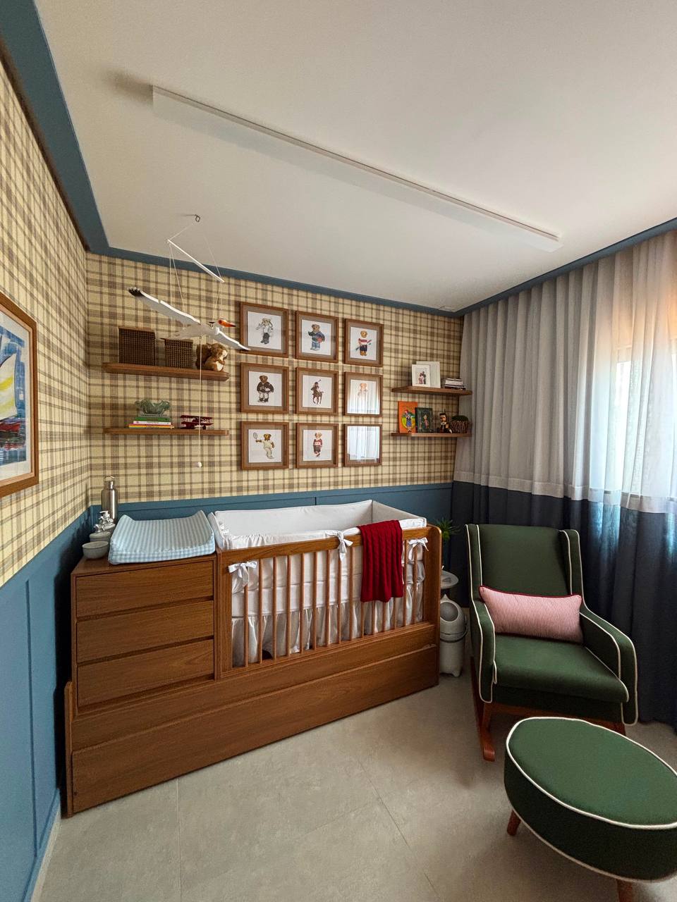 Quarto infantil com berço em madeira, papel de parede xadrez, boiserie azul e poltrona verde